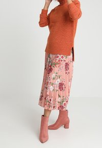 Maglione a maglia color ruggine con collo alto e spacchi laterali, abbinato a una gonna a pieghe floreale rosa e stivali alla caviglia rosa chiaro.