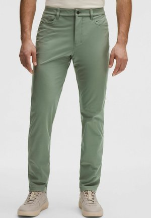 Mees, kes kannab kitsaid rohelist pükse ja heledaid beeže tossusid, seisab tavalise valge tausta ees.