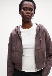 Brun zip-up hette med en teksturert finish, som har to frontlommer og en logo-patch, lagd over en hvit ribbet topp.