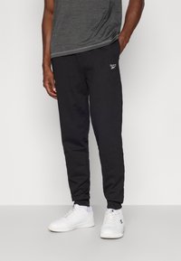 Čierne bavlnené tepláky s zúženými nohavicami a pružnými manžetami, na ľavej strane s malým bielym logom Reebok. Nosia sa s bielymi teniskami.
