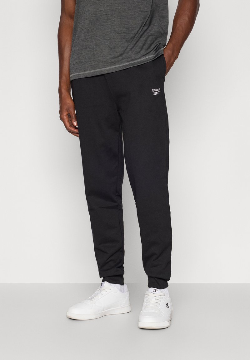 Čierne bavlnené tepláky s zúženými nohavicami a pružnými manžetami, na ľavej strane s malým bielym logom Reebok. Nosia sa s bielymi teniskami.
