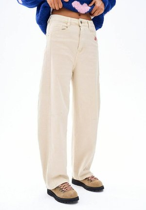 Osoba ubrana w beżowe szerokie spodnie, brązowe buty trekkingowe z różowymi sznurówkami oraz niebieski sweter z różowym serduszkiem, trzymająca rękami dół swetra.