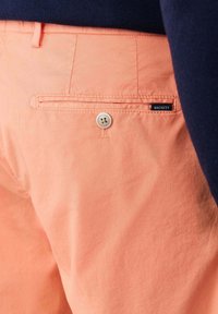 Hombre con pantalones de color coral con un bolsillo trasero abotonado y una pequeña etiqueta de la marca Hackett, combinado con una camiseta azul marino.