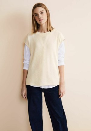 Cremfarbener, strukturierter Strickweste mit kurzen Ärmeln, die über einem weißen Langarmshirt getragen wird, kombiniert mit dunkelblauen Jeans.