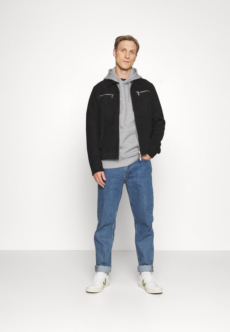 Hombre de pie con la mano en el bolsillo, que lleva chaqueta negra, sudadera con capucha gris, vaqueros azules y zapatillas blancas sobre un fondo blanco liso.