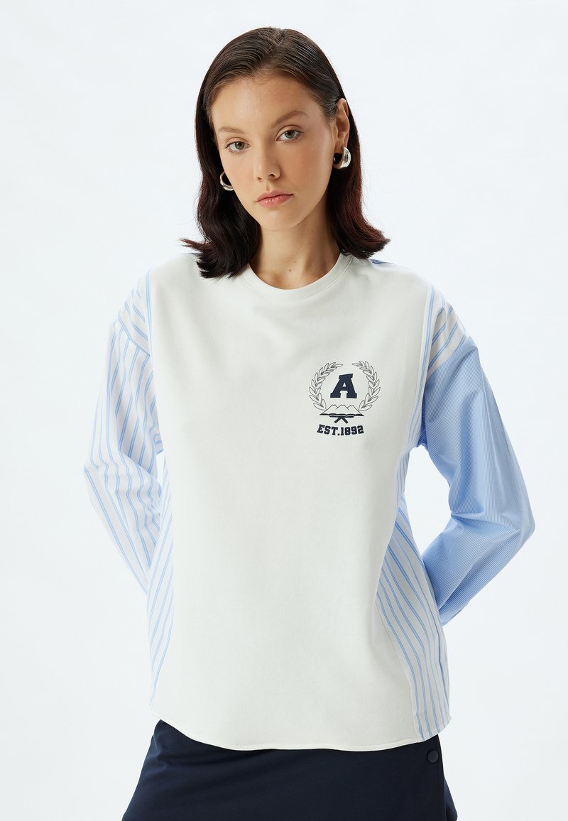 Sudadera blanca con mangas largas a rayas en azul claro y cuello redondo. Presenta un escudo con la letra "A" y la inscripción "EST. 1892".