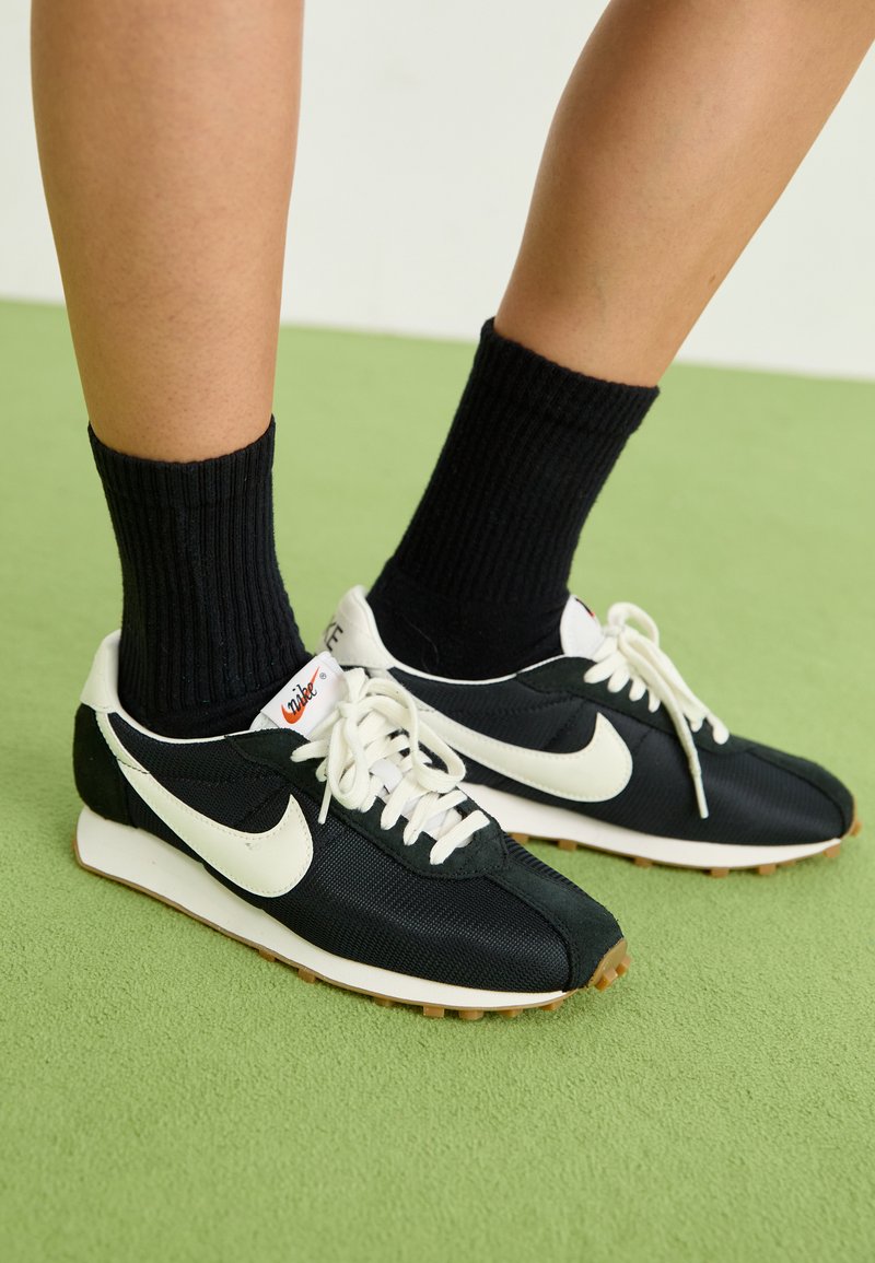 Crne Nike tenisice s bijelim akcentima, s gornjim dijelom od mrežastog materijala, vezanjem na vezice i gumeni potplat, uparen s crnim rebrastim čarapama.