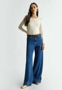 Maglietta beige a maniche lunghe abbinata a jeans blu a vita alta e gamba larga, con una cintura marrone. Scarpe bronze completano l'outfit.