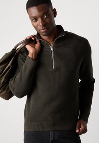 Suéter de canalé verde oscuro con un cuello de media cremallera. Lleva un bolso marrón, que presenta un diseño de punto texturizado y mangas largas.