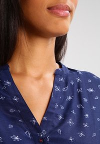 Blusa blu navy con stampe floreali e fogliari bianche, scollo a V e dettagli con bottoni. Tessuto morbido con una vestibilità comoda.