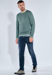 Hellgrüner Strickpullover mit rundem Ausschnitt, getragen über einem weißen Hemd, kombiniert mit dunkelblauen Jeans und grauen Turnschuhen. Einfaches, strukturiertes Design.