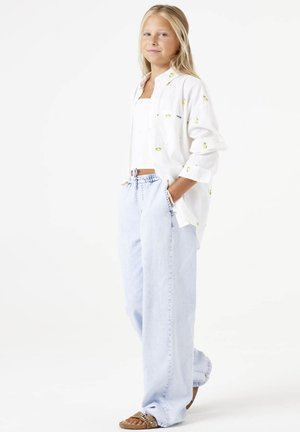 Camisa blanca de botón con bordado de frutas amarillas, combinada con jeans de pierna ancha azul claro y sandalias marrones con tachuelas decorativas.