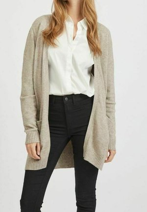 Kobieta w beżowym kardiganie z otwartym przodem, nosząca białą koszulę zapinaną na guziki oraz czarne spodnie z wysokim stanem, stojąca z rozluźnionymi rękami.