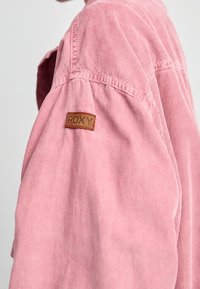 Manche de veste en velours côtelé rose avec une étiquette en cuir marron comportant l'inscription "ROXY" sur le haut du bras, vue recadrée sur un fond uni.