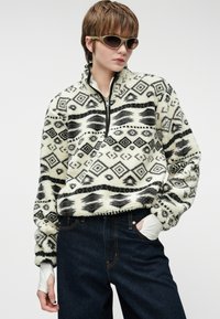 Fleece puloveris su juodai baltais geometriniais raštais, užtrauktuku apykaklėje ir plačiomis rankovėmis, derinamas su tamsiai mėlynomis džinsais ir balta марле.