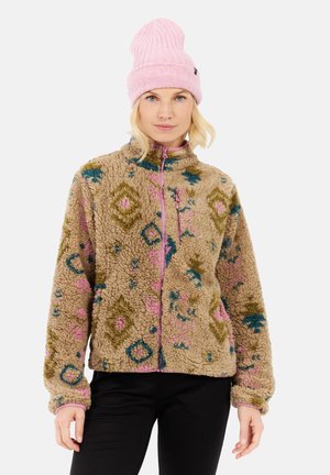 Donna che indossa una giacca in pile beige a fantasia con accenti rosa e blu e un berretto lavorato a maglia rosa, in piedi su uno sfondo bianco.