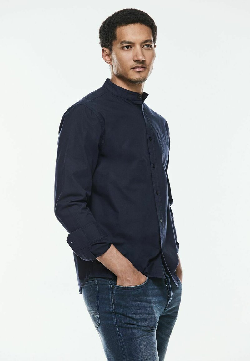 Camisa azul marino de botones con cuello mao, de manga larga y un bolsillo en el pecho, combinada con jeans oscuros de corte slim.