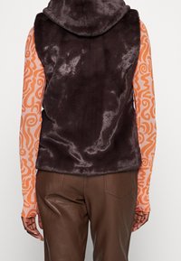 Braune Kunstfellweste mit Kapuze, die eine glatte Textur aufweist und keine Verschlüsse hat, getragen über einem langärmligen, orangefarbenen gemusterten Hemd.