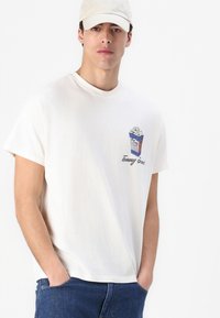 Hvit bomull t-skjorte med korte ermer, med et blått og hvitt popkorn-motiv på brystet og "Tommy Jeans"-tekst under.