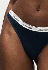 Άτομο με σκουρόχρωμο δέρμα που φορά μαύρα εσώρουχα Calvin Klein με λευκή μέση που φέρει επαναλαμβανόμενο λογότυπο της μάρκας, δείχνοντας το κάτω μέρος του κορμού και το επάνω μέρος των μηρών.