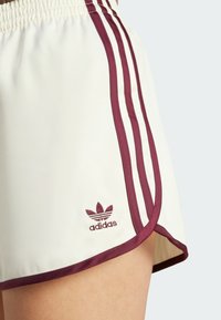 Krémové šortky s burgundskými postranními pruhy a lemem. Vybaveny vyšitým logem Adidas na dolním levém okraji. Lehký materiál s hladkou texturou.