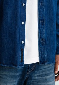 Man in een blauwe spijkerblouse over een witte T-shirt en blauwe spijkerbroek, hand rustend aan de zij, label zichtbaar aan de onderkant van de blouse.