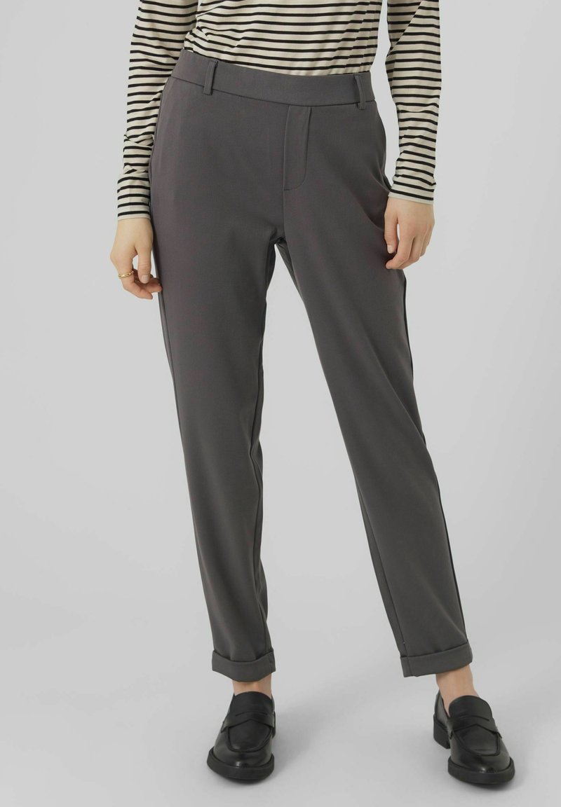 Personne portant un pantalon gris fuselé avec des ourlets retroussés, une chemise rayée noire et blanche à manches longues et des mocassins noirs, debout devant un fond uni.
