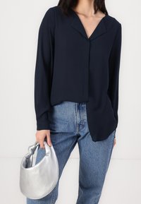 Blouse bleu marine à manches longues avec un col en V, associée à un jean taille haute bleu clair et un sac à main métallique argenté en forme de croissant.