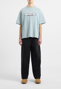Jasnoniebieski, oversize T-shirt z graficznym logo "AVX", w połączeniu z luźnymi czarnymi spodniami i beżowymi butami, stojący na neutralnym tle.