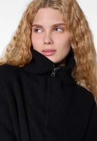 Pull à col montant noir à côtes avec demi-zip. Texture douce et coupe décontractée, mettant en avant un col volumineux et de subtils détails de couture.