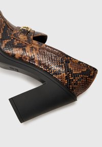 Καφέ τεχνητό δέρμα φιδιού με τακούνι, τετράγωνη μύτη και παχύ μαύρο μπλοκ τακούνι. Διαθέτει πλευρική ζώνη με χρυσή αγκράφα.