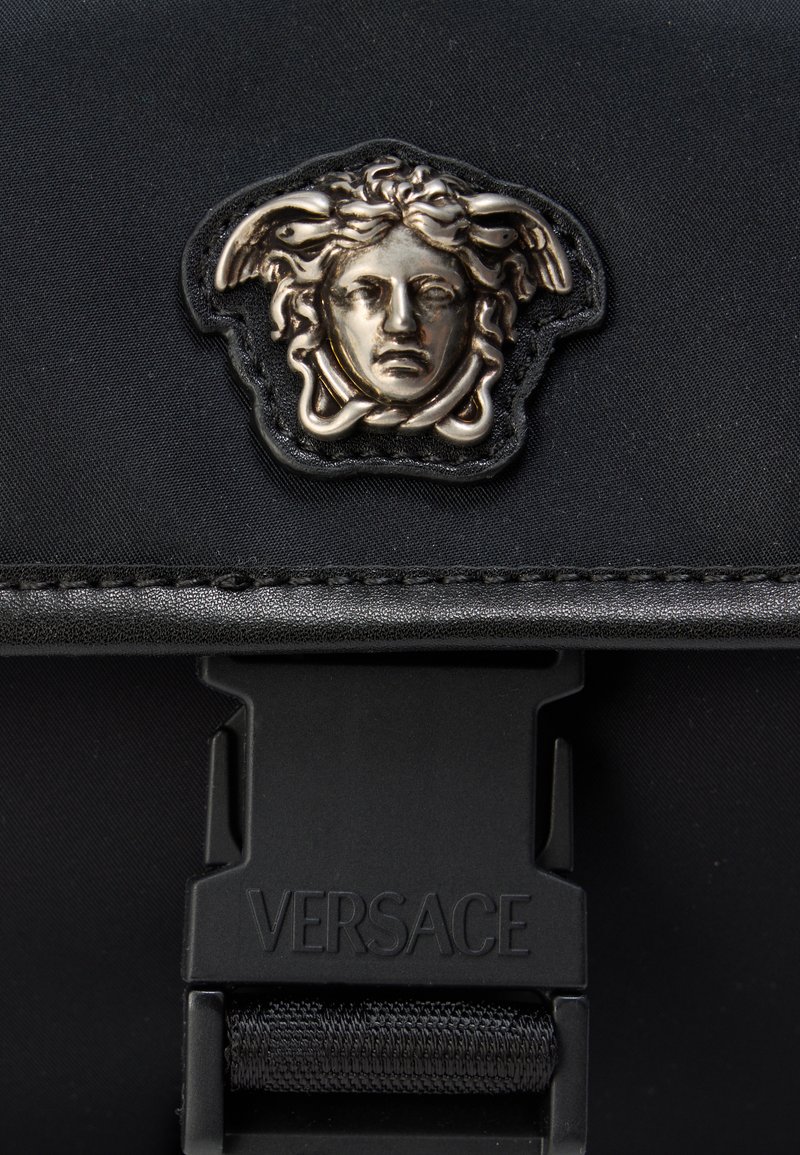Schwarze Nylon-Tasche mit einem erhabenen silbernen Medusa-Emblem und einem schwarzen verstellbaren Riemen mit einer Kunststoffschnalle, die mit "VERSACE" geprägt ist.