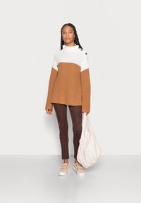 Pulôver tricotado bicolor com uma parte superior branca e uma parte inferior castanha, apresentando um gola alta e detalhes com botões laterais. Usado com calças castanhas e sapatos bege.