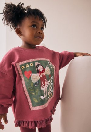 Jauns bērns ar cirtainiem matiem, valkājot rozā sporta krekliņu ar fejas ilustrāciju, stāv ar vienu roku uz gaišas krēsla.