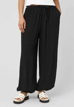 Femme portant un pantalon ample noir à cordon avec poches latérales et des sandales blanches à brides, debout devant un fond uni.