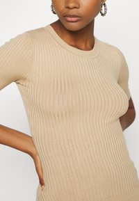 Beige ribbad stickad topp med korta ärmar och rund halsringning, med en mjuk textur och subtil elastiskitet för en figurnära silhuett.