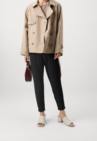  béžový dvouřadý kabát, černé kalhoty a krémové otevřené muly. Hladká tkanina s velkými knoflíky a uvolněným střihem. Drží burgundskou tašku.