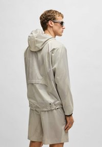 Lightweight, beige windbreaker with a hood, featuring a textured pattern and elastic cuffs. Paired with matching shorts, both have a relaxed fit.
