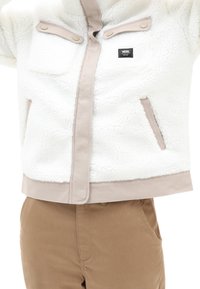 Hvid fleecejakke med glat beige kant, trykknapper og forlommer, sammen med lysebrune bukser.