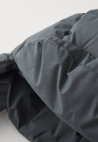 Dunkelgraue Daunenjacke mit einer vom Steppmuster geprägten Struktur, glänzender Oberfläche und auffälligen Nähdetails. Weiches, isoliertes Material sichtbar.