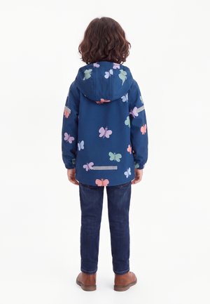 Barn iført en blå jakke med flerfarvede sommerfugleprint, mørke jeans og brune sko, stående med ryggen mod på hvid baggrund.