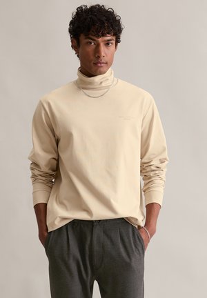 Beige Langarm-Turtleneck-Shirt aus weichem Stoff, mit einem dezenten, in heller Farbe gedruckten Logo, kombiniert mit grauen, gefalteten Hosen.