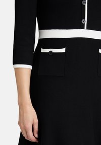 Robe noire avec accents blancs, devant boutonné, et poignets côtelés avec poches. Tissu en maille doux et texturé avec une coupe ajustée.