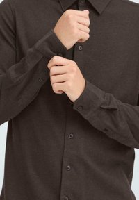 Brun langermet skjorte med krage som kan knappes ned, med mørke knapper og en myk tekstur, fremhevet av oppbrettede ermer.