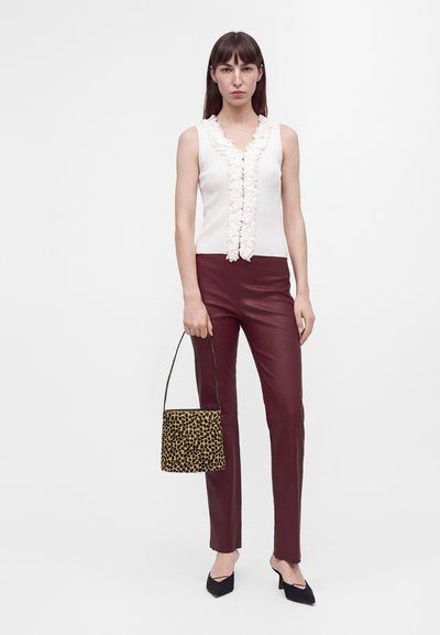 Femme portant un haut blanc sans manches à volants, pantalon en cuir bordeaux, talons noirs pointus, tenant un sac à main imprimé léopard sur fond blanc.
