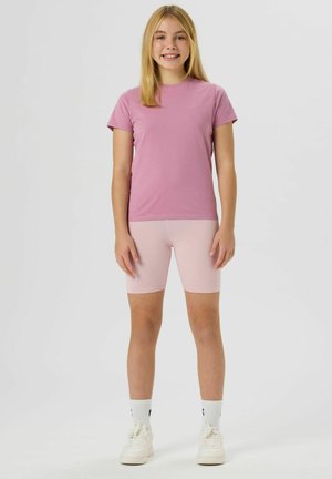 Fille aux cheveux blonds debout, souriante, portant un t-shirt rose, un short rose clair, des chaussettes blanches et des baskets blanches sur un fond uni.