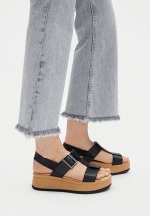 Füße in schwarzen Leder-Plattformsandalen mit großen Schnallen, kombiniert mit hellblauen Jeans mit ausgefranstem Saum.