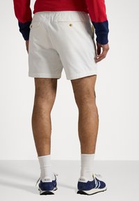 Muž oblečený v bílých casual šortkách s knoflíkovými zadními kapsami, bílých crew ponožkách a tmavomodrých teniskách s bílými tkaničkami stojí na jednobarevném pozadí.