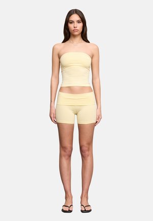 Donna in piedi rivolta verso l'obiettivo, indossa un top a fascia giallo pallido e pantaloncini coordinati con infradito nere su uno sfondo chiaro semplice.