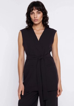 Kobieta o ciemnych falowanych włosach, ubrana w bezrękawnik z paskiem w kolorze czarnym oraz dopasowane czarne spodnie, stoi na tle jednolitego białego tła.